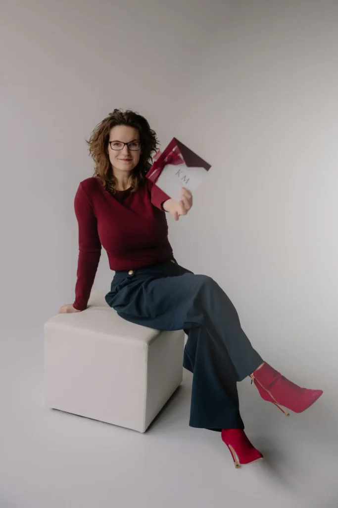 Aneta Różycka ubrana w kolory swojej marki czyli mordo i szarość, trzymająca jedno ze swoich zaproszeń w kolorze bordo