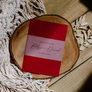 Bordowa koperta na zaproszenie, zaprezentowana na kawałku drewnianego pnia, w tle bielony ruskus